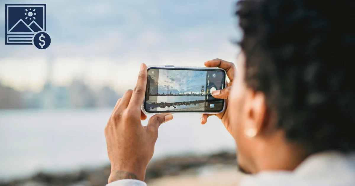 A person holding a smartphone horizontally, taking a photo of a cityscape across a body of water—perfect for anyone starting a stock photography side hustle. The phone’s screen captures the scene, with blurred buildings in the background.