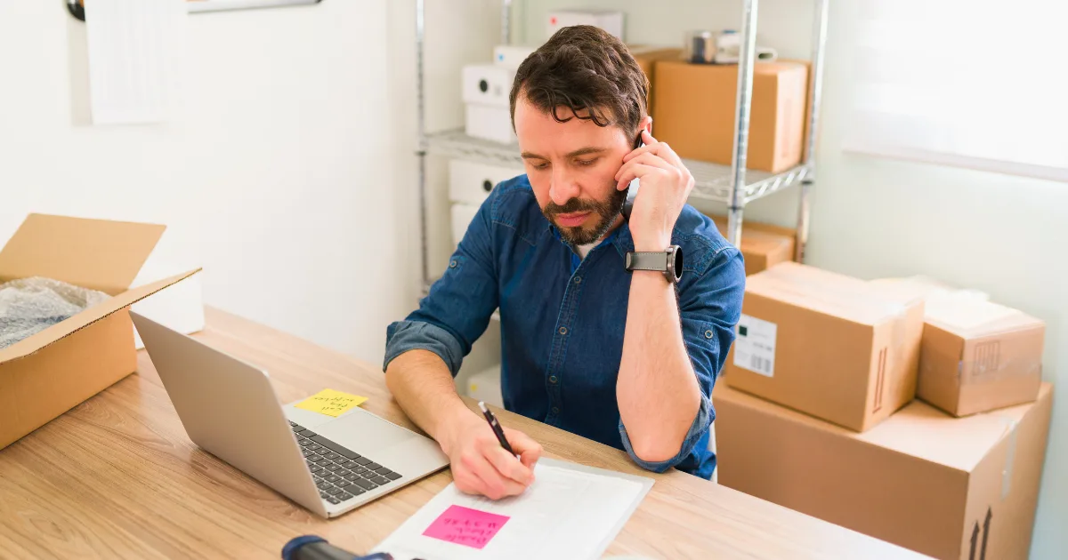 A man sits at a desk with a laptop and cardboard boxes, talking on the phone—likely managing a supplier relationship—and writing notes on paper in a room with shelves and bright light.
