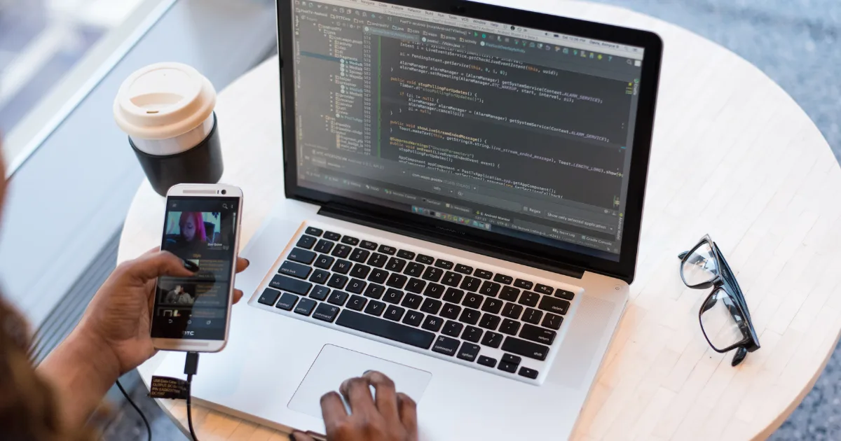 A person works on a laptop displaying code, likely focusing on a web development side hustle, while holding a smartphone. A pair of glasses and a takeaway coffee cup rest on the round wooden table.