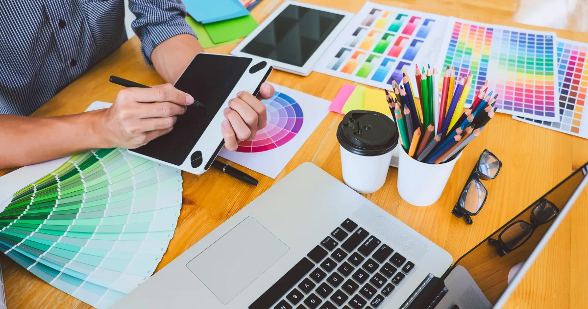 A person uses a graphic tablet at a desk with a laptop, color swatches, pencils, coffee cup, and digital tablets—exploring graphic design tools for non-designers amid colorful design materials and creative resources.