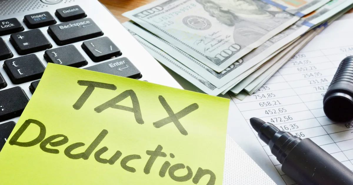 A sticky note with TAX Deduction written on it sits on a laptop keyboard next to a stack of US $100 bills, a black marker, and a financial document—perfect for tracking home office tax deductions.